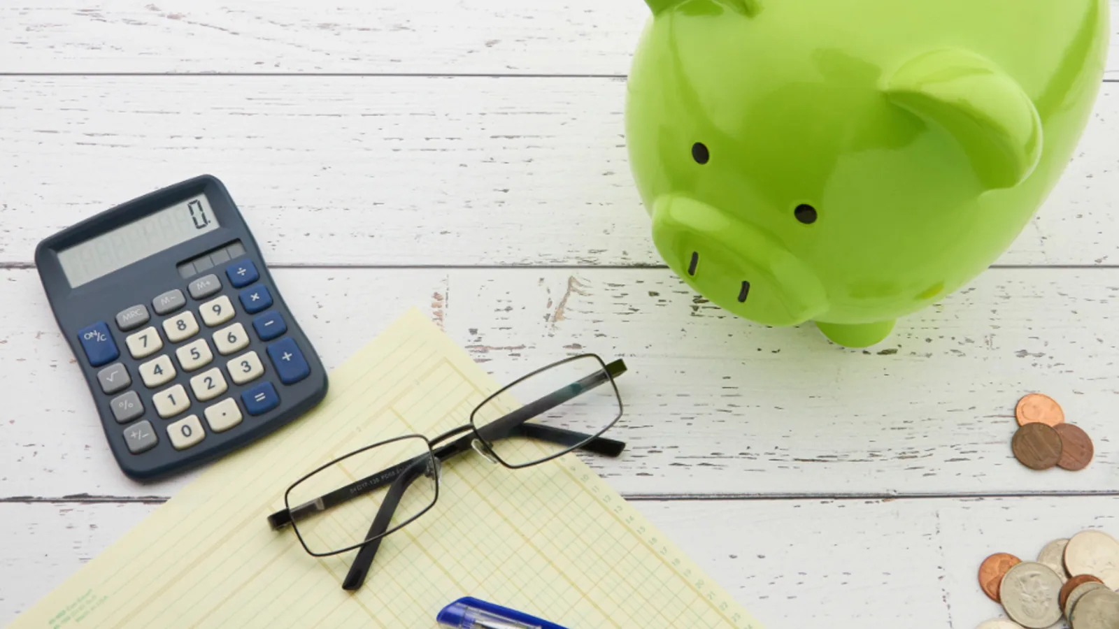 Green piggy bank beside a calculator, glasses and a notepad
